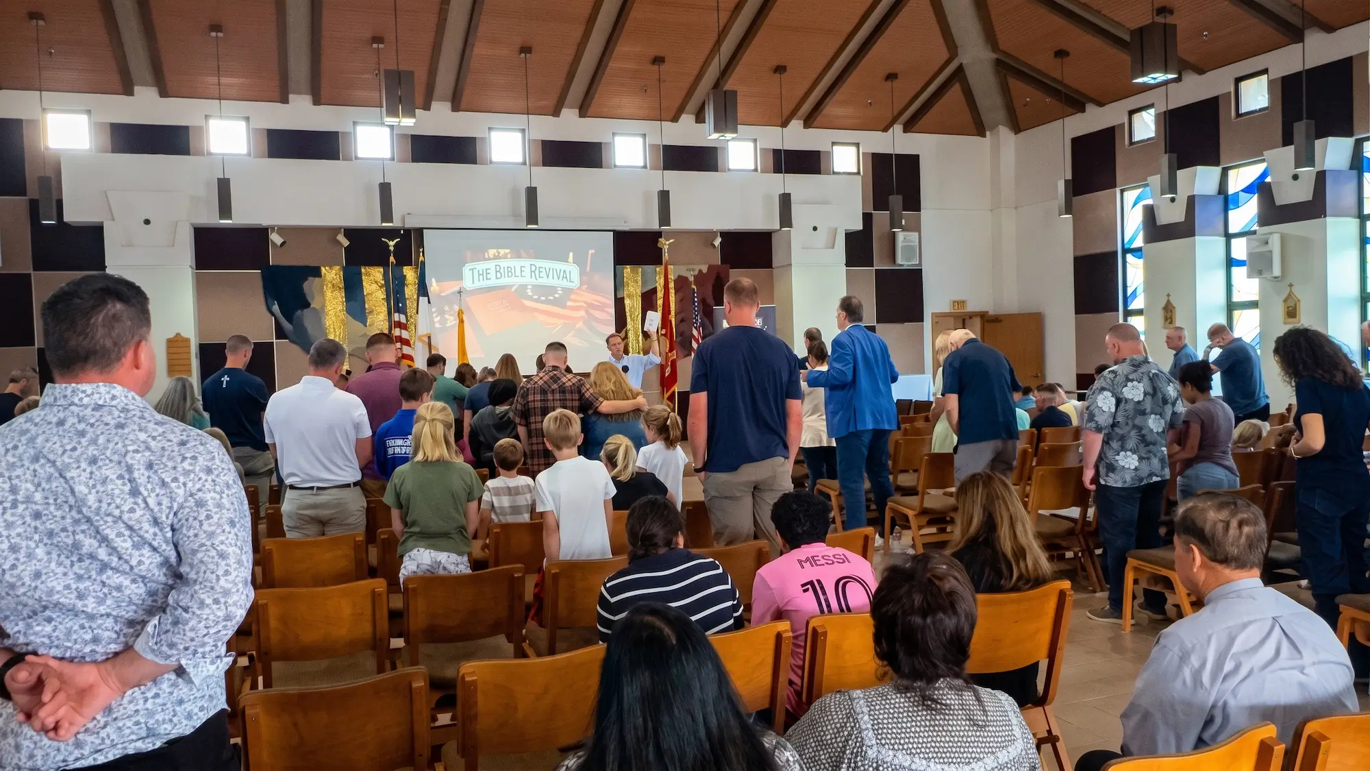 Families standing together at a Bible Revival assembly as Kirk Cameron holds up a Bible on stage