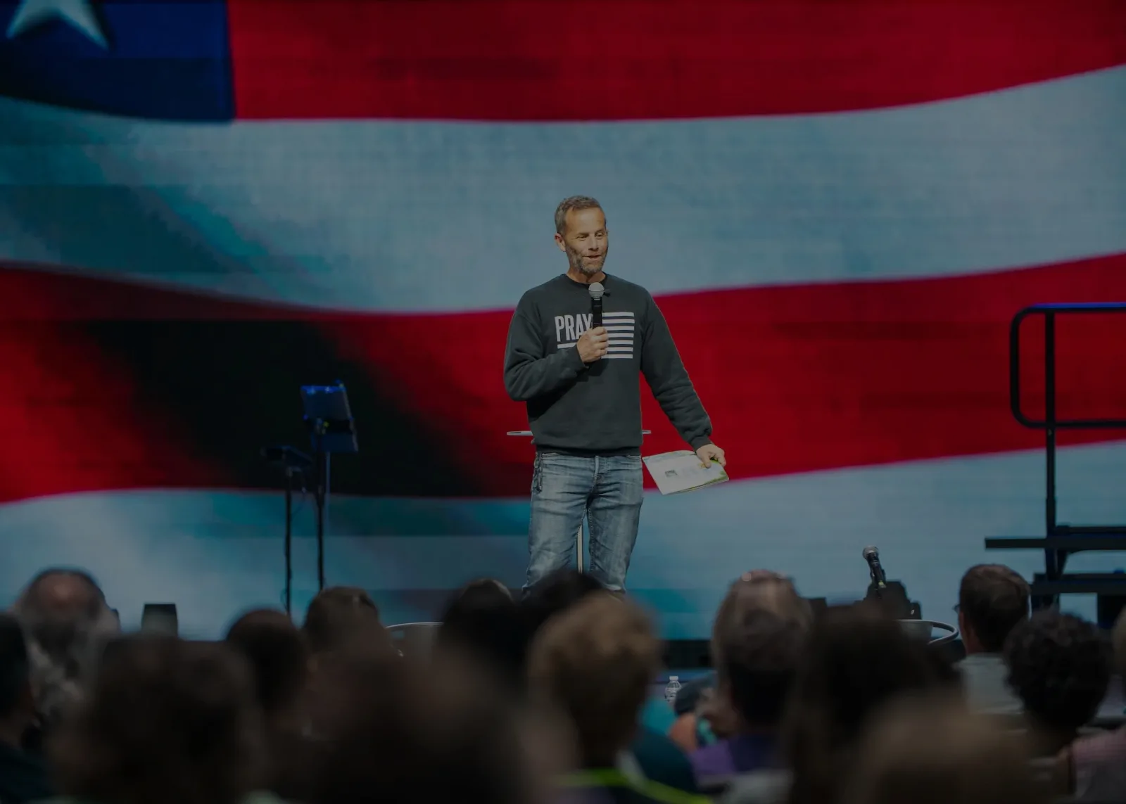 Kirk Cameron reading to a packed auditorium of families in front of an American flag backdrop